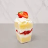 3 leches en vasito. Un vasito cuadrado transparente que muestra un postre de tres leches estratificado. Comienza con una base de pan esponja claro, seguida de una capa de crema, abundantes fresas frescas picadas, y otra capa de pan. Remata con un generoso copo de crema batida y media fresa fresca en la parte superior.