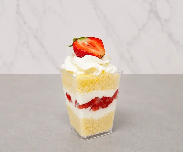 3 leches en vasito. Un vasito cuadrado transparente que muestra un postre de tres leches estratificado. Comienza con una base de pan esponja claro, seguida de una capa de crema, abundantes fresas frescas picadas, y otra capa de pan. Remata con un generoso copo de crema batida y media fresa fresca en la parte superior.