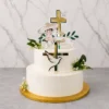 Tierno pastel de bautizo de dos pisos en color blanco liso. Resalta una gran cruz de acrílico dorado que atraviesa los pisos, con la figura de una paloma blanca. Está adornado con toques florales de follaje verde y flores nube, rematado con un adorno de un niño con traje de charro y un listón con el nombre "Paquito". Castillo's Cake Shop.