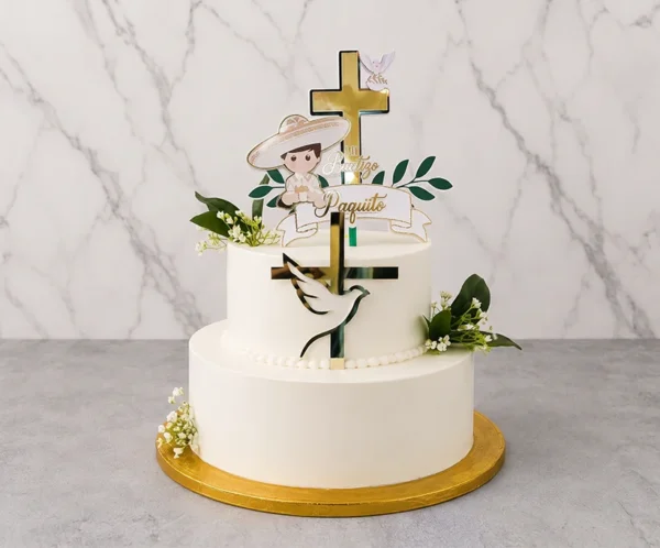 Tierno pastel de bautizo de dos pisos en color blanco liso. Resalta una gran cruz de acrílico dorado que atraviesa los pisos, con la figura de una paloma blanca. Está adornado con toques florales de follaje verde y flores nube, rematado con un adorno de un niño con traje de charro y un listón con el nombre "Paquito". Castillo's Cake Shop.