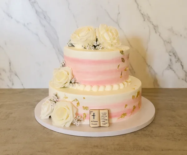 Delicado pastel de dos pisos con un suave diseño de líneas difuminadas en tonos blanco y rosa pastel. Está hermosamente decorado con destellos de hoja de oro, abundantes rosas blancas con pequeñas flores nube y un sutil librito de fondant en la base que dice "Mi Primera Comunión". Castillo's Cake Shop.