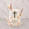 Moderno pastel de dos pisos estilo boho con cobertura blanca lisa. En el piso superior destaca una cruz negra con bordes dorados, y en el inferior el nombre "Leylani" en acrílico dorado. Está adornado con flores de glaseado en tonos blanco y palo de rosa, complementado con hermosas espigas y pasto pampa seco. Castillo's Cake Shop.