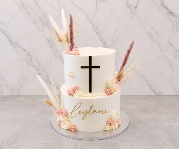 Moderno pastel de dos pisos estilo boho con cobertura blanca lisa. En el piso superior destaca una cruz negra con bordes dorados, y en el inferior el nombre "Leylani" en acrílico dorado. Está adornado con flores de glaseado en tonos blanco y palo de rosa, complementado con hermosas espigas y pasto pampa seco. Castillo's Cake Shop.