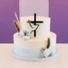 Sofisticado pastel de dos pisos con un suave degradado en acuarela color azul claro en la base. Su diseño celestial incluye una cruz negra con una paloma blanca en acrílico, macarrones blancos, estrellitas de glaseado azul, hojitas de fondant y sutiles espigas secas en tonos tierra y azulados. Castillo's Cake Shop.