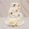 Bodas1 Imponente pastel de bodas de cuatro pisos en color blanco, decorado con un elaborado estilo clásico (Lambeth) que incluye múltiples guirnaldas, cortinajes, puntos y bordes festoneados. Cuenta con grandes flores en tonos crema y amarillo pálido, además de las letras "B", "J" e "Y" colocadas en los tres pisos superiores.