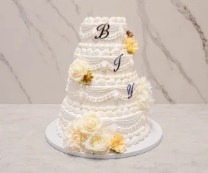 Bodas1 Imponente pastel de bodas de cuatro pisos en color blanco, decorado con un elaborado estilo clásico (Lambeth) que incluye múltiples guirnaldas, cortinajes, puntos y bordes festoneados. Cuenta con grandes flores en tonos crema y amarillo pálido, además de las letras "B", "J" e "Y" colocadas en los tres pisos superiores.