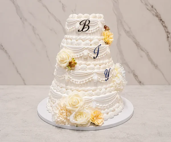 Bodas1 Imponente pastel de bodas de cuatro pisos en color blanco, decorado con un elaborado estilo clásico (Lambeth) que incluye múltiples guirnaldas, cortinajes, puntos y bordes festoneados. Cuenta con grandes flores en tonos crema y amarillo pálido, además de las letras "B", "J" e "Y" colocadas en los tres pisos superiores.