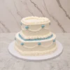Bodas11 Romántico pastel de bodas de dos pisos, ambos con forma de corazón. Presenta una base de glaseado blanco liso, decorada al estilo Lambeth con intrincados bordes de concha y guirnaldas punteadas. Destacan delicados detalles en color azul pálido, incluyendo el borde inferior del piso superior y pequeños corazones de glaseado a lo largo de las guirnaldas.