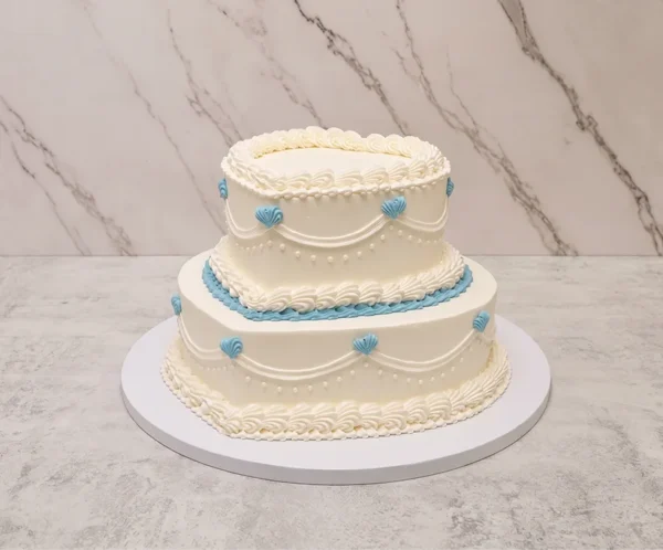 Bodas11 Romántico pastel de bodas de dos pisos, ambos con forma de corazón. Presenta una base de glaseado blanco liso, decorada al estilo Lambeth con intrincados bordes de concha y guirnaldas punteadas. Destacan delicados detalles en color azul pálido, incluyendo el borde inferior del piso superior y pequeños corazones de glaseado a lo largo de las guirnaldas.