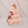 Bodas12 Imponente pastel de bodas de cuatro pisos con cobertura lisa en un suave tono rosa rubor o melocotón claro. Cada piso está finamente adornado con aplicaciones de volutas y florituras de fondant en un tono beige o dorado apagado. Presenta un exquisito arreglo floral en tonos complementarios que adorna la corona y desciende por un costado hasta la base.