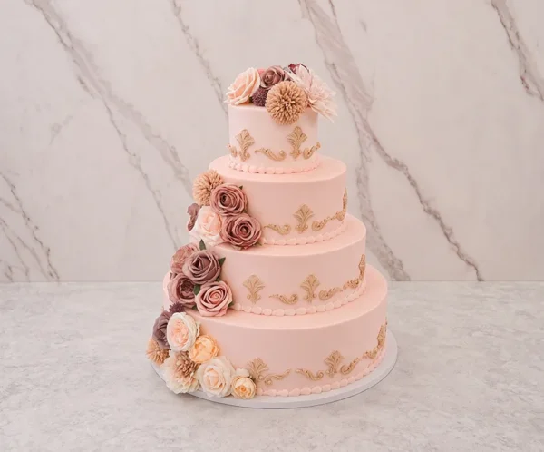 Bodas12 Imponente pastel de bodas de cuatro pisos con cobertura lisa en un suave tono rosa rubor o melocotón claro. Cada piso está finamente adornado con aplicaciones de volutas y florituras de fondant en un tono beige o dorado apagado. Presenta un exquisito arreglo floral en tonos complementarios que adorna la corona y desciende por un costado hasta la base.
