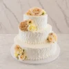 Bodas2 Pastel de bodas de tres pisos con glaseado blanco que destaca por un diseño de textura rústica con ondas pronunciadas y espatuladas. Está decorado con arreglos florales en tonos melocotón, crema y amarillo suave, dispuestos estratégicamente en la parte superior, el centro y la base.