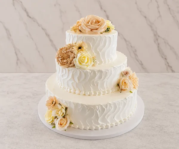 Bodas2 Pastel de bodas de tres pisos con glaseado blanco que destaca por un diseño de textura rústica con ondas pronunciadas y espatuladas. Está decorado con arreglos florales en tonos melocotón, crema y amarillo suave, dispuestos estratégicamente en la parte superior, el centro y la base.