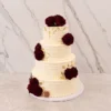 Bodas6 Pastel de bodas de cuatro pisos con cobertura color crema y sutiles líneas horizontales texturizadas. Está decorado elegantemente con una cascada asimétrica de rosas en color rojo vino profundo y delicados toques de hoja de oro esparcidos por la superficie de los niveles.