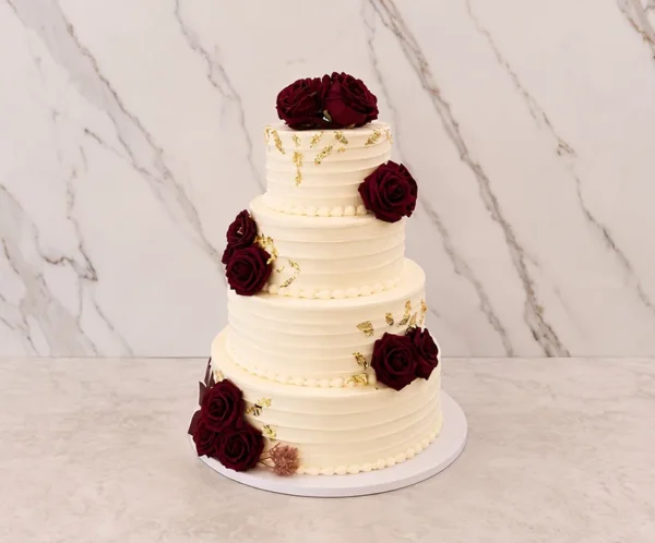 Bodas6 Pastel de bodas de cuatro pisos con cobertura color crema y sutiles líneas horizontales texturizadas. Está decorado elegantemente con una cascada asimétrica de rosas en color rojo vino profundo y delicados toques de hoja de oro esparcidos por la superficie de los niveles.