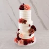 Bodas7 Elegante pastel de bodas de tres pisos con glaseado blanco de textura acanalada horizontal. El diseño resalta por un arreglo floral asimétrico en tonos rojo vino, rosa viejo y palo de rosa que trepa desde la base hasta la corona, con hojas oscuras para darle profundidad. La base del piso inferior está envuelta en un sutil listón rosa metálico.