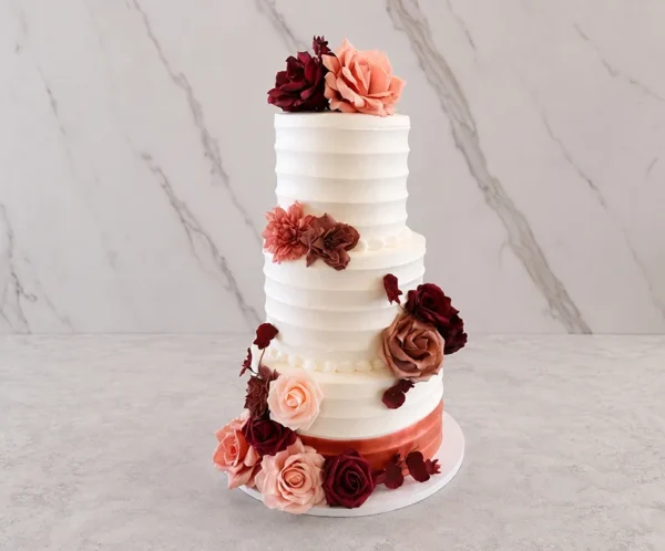 Bodas7 Elegante pastel de bodas de tres pisos con glaseado blanco de textura acanalada horizontal. El diseño resalta por un arreglo floral asimétrico en tonos rojo vino, rosa viejo y palo de rosa que trepa desde la base hasta la corona, con hojas oscuras para darle profundidad. La base del piso inferior está envuelta en un sutil listón rosa metálico.