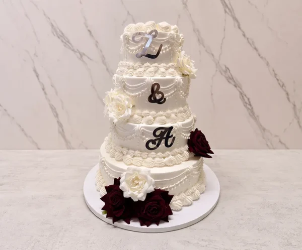 Bodas8 Sofisticado pastel de bodas de cuatro pisos cubierto con fondant blanco. Está elegantemente decorado con guirnaldas y bordes estilo Lambeth. Presenta las iniciales "L & A" en letras acrílicas de espejo plateado oscuro. El contraste se logra con hermosas rosas en tono rojo vino en la base y el piso inferior, complementadas con rosas blancas.