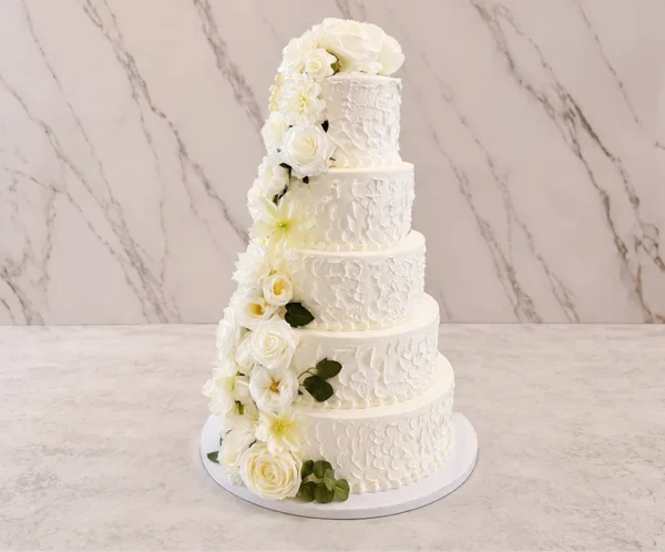 Bodas9 Majestuoso pastel de bodas de cinco pisos con glaseado blanco que luce un patrón espatulado rústico en toda la superficie. El diseño destaca por una hermosa y tupida cascada de flores blancas, incluyendo rosas y lirios, acompañadas de follaje verde oscuro que fluye desde la parte superior hasta la base.