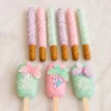 Conjunto de postres personalizados en armoniosos tonos pastel lila, rosa y verde menta. En la parte superior, se muestran seis palitos de pretzel cubiertos de chocolate (dos de cada color) y espolvoreados con chispas blancas. En la parte inferior, tres paletas tipo Magnum (cakesicles) decoradas con líneas finas, racimos de chispas de colores y grandes mariposas comestibles translúcidas posadas en la superficie.