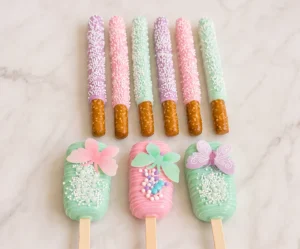 Conjunto de postres personalizados en armoniosos tonos pastel lila, rosa y verde menta. En la parte superior, se muestran seis palitos de pretzel cubiertos de chocolate (dos de cada color) y espolvoreados con chispas blancas. En la parte inferior, tres paletas tipo Magnum (cakesicles) decoradas con líneas finas, racimos de chispas de colores y grandes mariposas comestibles translúcidas posadas en la superficie.