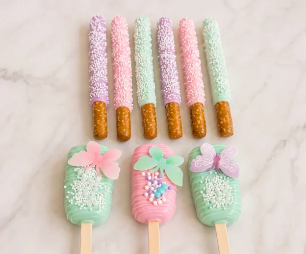 Conjunto de postres personalizados en armoniosos tonos pastel lila, rosa y verde menta. En la parte superior, se muestran seis palitos de pretzel cubiertos de chocolate (dos de cada color) y espolvoreados con chispas blancas. En la parte inferior, tres paletas tipo Magnum (cakesicles) decoradas con líneas finas, racimos de chispas de colores y grandes mariposas comestibles translúcidas posadas en la superficie.