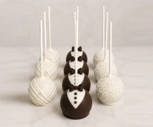 Elegantes cake pops con temática formal o de boda organizadas en tres filas. La primera fila está cubierta de chocolate blanco con un diseño de líneas diagonales texturizadas. La fila central presenta un diseño de esmoquin con chocolate oscuro, pechera blanca, botones y un pequeño moño. La tercera fila está completamente cubierta de chocolate blanco y adornada con diminutas chispas redondas.