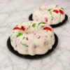 Dos refrescantes gelatinas de mosaico presentadas en forma de rosca tradicional. Destacan por su base cremosa de leche blanca que encapsula vibrantes cubos de gelatina translúcida en colores rojo y verde. Castillo's Cake Shop.