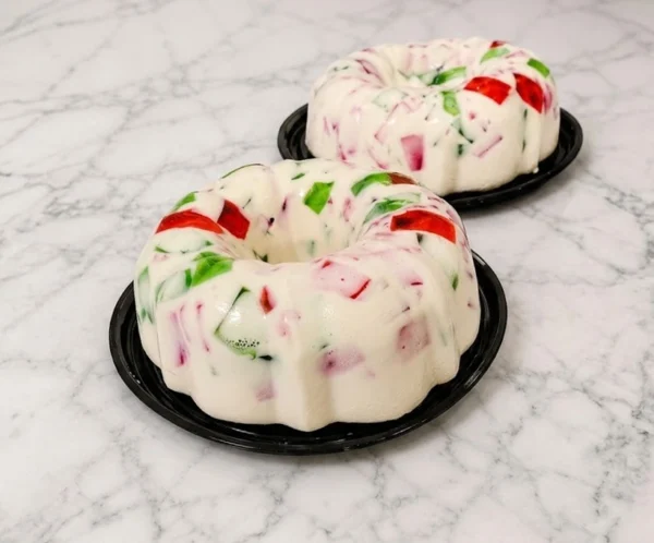 Dos refrescantes gelatinas de mosaico presentadas en forma de rosca tradicional. Destacan por su base cremosa de leche blanca que encapsula vibrantes cubos de gelatina translúcida en colores rojo y verde. Castillo's Cake Shop.