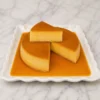 Tradicional flan napolitano redondo, liso y cremoso, bañado en un rico jarabe de caramelo. Se presenta en un plato blanco de bordes ondulados con una rebanada cortada y delicadamente superpuesta para mostrar su textura perfecta. Castillo's Cake Shop.