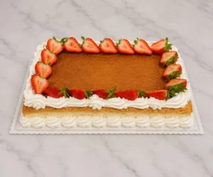Atractivo pastel rectangular tipo plancha con una superficie cubierta de una suave capa de cajeta o dulce de leche. Está enmarcado por tupidos bordes de crema batida blanca y decorado con mitades de fresas frescas alrededor de todo el perímetro. Castillo's Cake Shop.