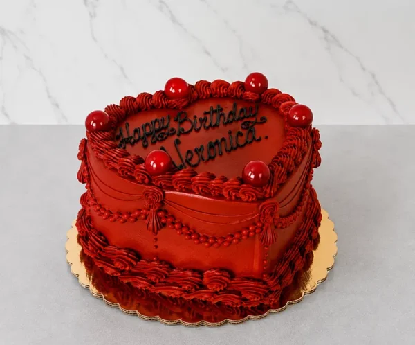 Pastel en forma de corazón cubierto completamente con un intenso glaseado color rojo oscuro. Su diseño clásico incluye detalles de puntos, drapeados elaborados, esferas rojas brillantes decorativas y el mensaje "Happy Birthday Veronica." escrito en negro. Castillo's Cake Shop.