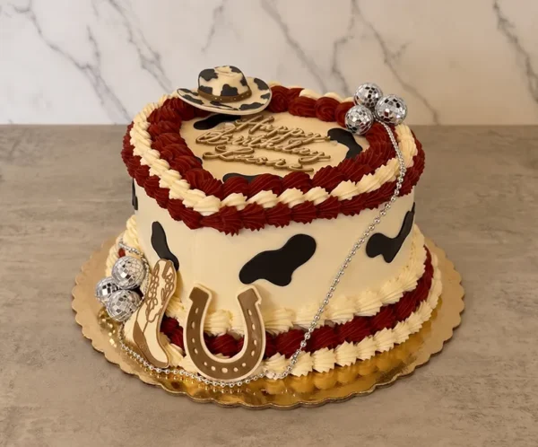 Original pastel en forma de corazón con temática vaquera y disco. Tiene una base blanca con manchas de vaca negras, contrastada con gruesos bordes trenzados en glaseado color vino y crema. Está adornado con mini bolas de discoteca, una cadena plateada, figuras decorativas de un sombrero, una herradura y una bota vaquera, rematado con un letrero dorado de "Happy Birthday Leon". Castillo's Cake Shop.