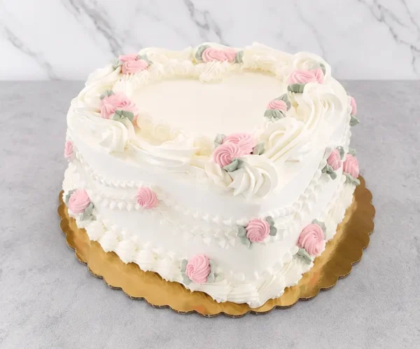 Clásico y romántico pastel en forma de corazón con cobertura blanca pura. Está hermosamente decorado con volutas, guirnaldas y bordes de glaseado blanco estilo vintage, acentuado a lo largo de su diseño con delicadas rosas de glaseado rosa pastel y sutiles hojas verdes. Castillo's Cake Shop.