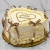 Pastel de aniversario en forma de corazón con glaseado color crema y una intrincada decoración estilo Lambeth. Resalta por sus elegantes bordes, perlas doradas comestibles, un corazón formado por perlas doradas con el mensaje "Happy Anniversary" en el centro, y hermosos lazos de cinta satinada en tono café. Castillo's Cake Shop.