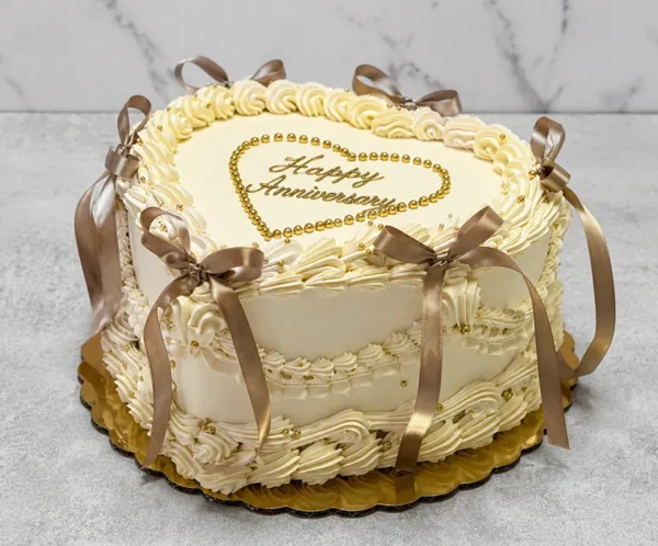 Pastel de aniversario en forma de corazón con glaseado color crema y una intrincada decoración estilo Lambeth. Resalta por sus elegantes bordes, perlas doradas comestibles, un corazón formado por perlas doradas con el mensaje "Happy Anniversary" en el centro, y hermosos lazos de cinta satinada en tono café. Castillo's Cake Shop.