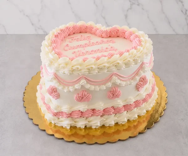 Pastel en forma de corazón con una base de glaseado blanco y delicados detalles estilo vintage en color rosa, que incluyen elegantes guirnaldas y bordes de concha. En la superficie lleva el mensaje "Feliz Cumpleaños Veronica" en caligrafía de glaseado rosa. Castillo's Cake Shop.