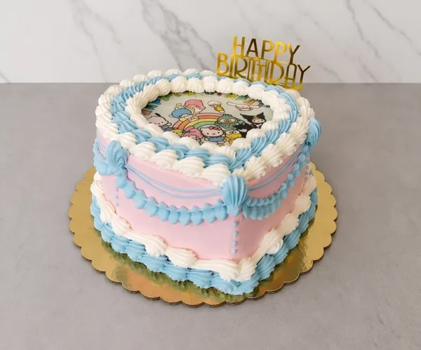 Pastel en forma de corazón con base color rosa pastel, decorado con elaborados bordes de glaseado entrelazado en tonos azul claro y blanco estilo Lambeth. En el centro superior lleva una colorida imagen comestible con personajes de Sanrio sobre un arcoíris y está coronado con un letrero dorado de "HAPPY BIRTHDAY". Castillo's Cake Shop.