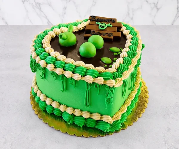 Divertido pastel en forma de corazón con temática de Shrek. Cuenta con una base de glaseado verde vibrante y un diseño de "pantano" en la superficie con un charco de chocolate oscuro y esferas verdes tridimensionales simulando burbujas. Está decorado con gruesos bordes de glaseado en verde y crema, detalles de enredaderas colgantes y un letrero que dice "Beware OGRO". Castillo's Cake Shop.