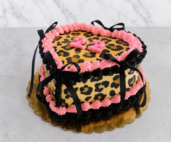 Llamativo pastel en forma de corazón con un audaz diseño de estampado de leopardo en la superficie y los costados. Los bordes están decorados con rosetones de glaseado en color rosa y negro profundo, acentuados con elegantes lazos de cinta negra satinada y el número "22" en color rosa en el centro. Castillo's Cake Shop.