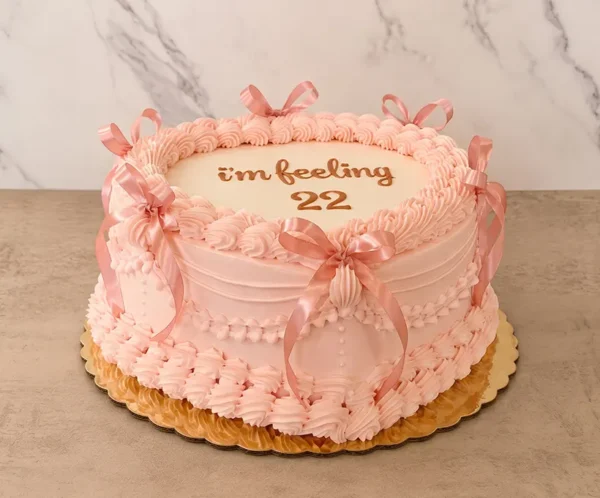Elegante y delicado pastel en forma de corazón cubierto en un tono rosa suave. Destaca por su decoración estilo Lambeth con intrincados bordes, guirnaldas acanaladas, detalles de perlas de glaseado y hermosos lazos de cinta rosa. En el centro de la superficie luce el mensaje "i'm feeling 22" en sutiles letras doradas. Castillo's Cake Shop.