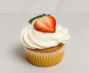 CupCake2 Tradicional cupcake de vainilla cubierto con un perfecto y cremoso remolino de glaseado blanco, decorado de forma fresca con la mitad de una jugosa fresa natural en la cima. Castillo's Cake Shop.
