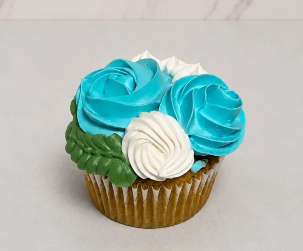 Cupcake clásico decorado con un hermoso arreglo floral de glaseado, que incluye dos vibrantes rosas color azul turquesa, flores blancas y hojas verdes entrelazadas. Castillo's Cake Shop.