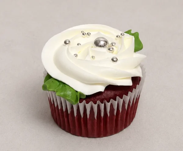 Elegante cupcake red velvet coronado con una gran rosa de glaseado blanco. Está finamente adornado con perlas plateadas comestibles de diferentes tamaños y una delicada hoja verde en la base. Castillo's Cake Shop.