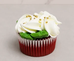 Esponjoso cupcake red velvet con un generoso remolino de glaseado blanco, acentuado con elegantes perlas doradas esparcidas en la superficie y hojitas de glaseado verde. Castillo's Cake Shop.