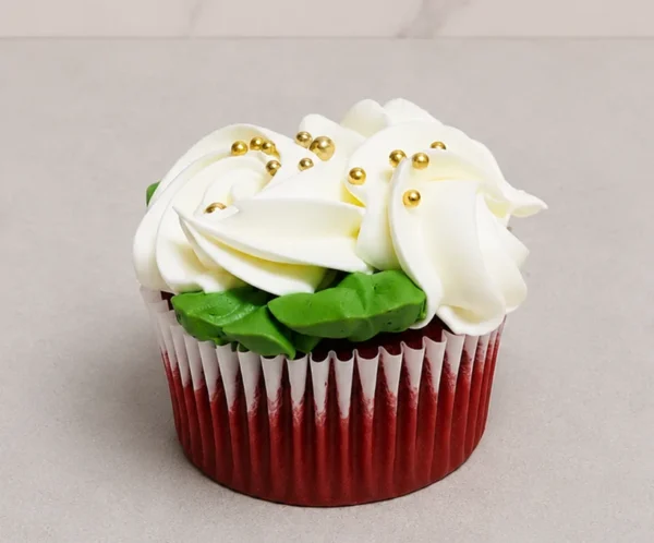 Esponjoso cupcake red velvet con un generoso remolino de glaseado blanco, acentuado con elegantes perlas doradas esparcidas en la superficie y hojitas de glaseado verde. Castillo's Cake Shop.