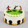 Pastel redondo con temática de fútbol. Los laterales están decorados con hexágonos blancos y negros de fondant simulando la textura de un balón, mientras que la parte superior representa una cancha verde lisa con bordes de crema blanca. Incluye una portería y tres figuras plásticas de jugadores en acción. Castillo's Cake Shop.