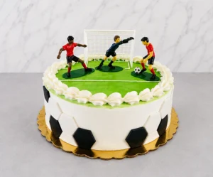 Pastel redondo con temática de fútbol. Los laterales están decorados con hexágonos blancos y negros de fondant simulando la textura de un balón, mientras que la parte superior representa una cancha verde lisa con bordes de crema blanca. Incluye una portería y tres figuras plásticas de jugadores en acción. Castillo's Cake Shop.