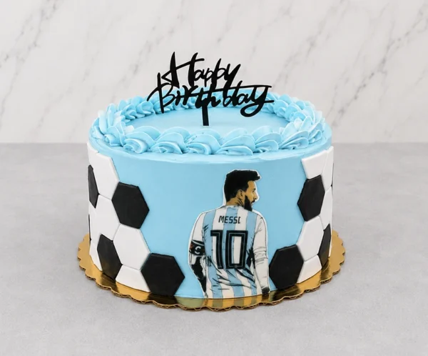 Pastel redondo cubierto de glaseado azul celeste con bordes acanalados en la parte superior. Los costados tienen un diseño parcial de balón de fútbol con hexágonos de fondant. Al frente destaca una imagen de Lionel Messi de espaldas con la camiseta número 10 de Argentina, rematado con un letrero negro de "Happy Birthday". Castillo's Cake Shop.