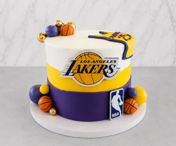 Elegante pastel cilíndrico con los colores de Los Angeles Lakers: franjas gruesas en blanco, amarillo y morado. En el centro lleva el logotipo de los Lakers y el escudo de la NBA en la base. Está decorado con esferas en morado, amarillo, dorado y pequeñas pelotas de básquetbol, además del detalle de la camiseta con el número 23 sobre el borde superior. Castillo's Cake Shop.